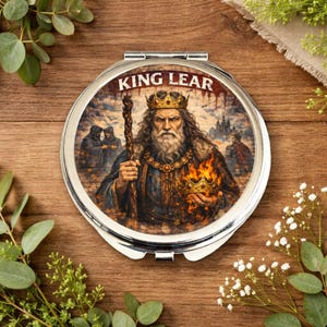 Puede incluir: Un espejo compacto redondo y plateado con las palabras "KING LEAR" en la parte superior. El diseño del espejo presenta una ilustración detallada del Rey Lear, sosteniendo un bastón y una corona en llamas. El fondo muestra figuras y un castillo.