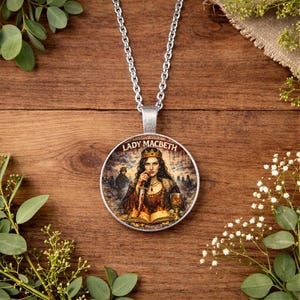 Peut inclure: Un collier pendentif argenté avec un pendentif rond portant l'inscription "LADY MACBETH". Le pendentif représente une femme couronnée tenant un poignard, avec un livre et un gobelet. Le collier est posé sur une surface en bois.