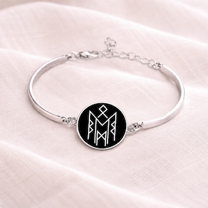 May include: Silver-toned bangle bracelet featuring a circular black pendant with a white geometric design. The bracelet has a simple, elegant design with an adjustable chain closure. The pendant's design is a stylized symbol.