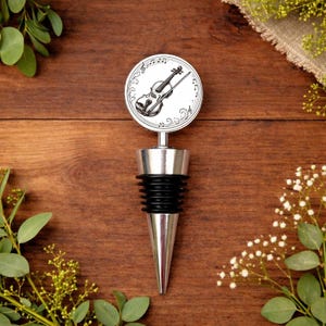 May include: A silver wine bottle stopper with a black rubber seal and a conical tip. The top has a circular design with a violin illustration and musical notes. The background is a wooden surface with greenery.