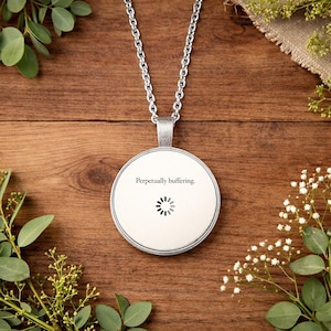 Puede incluir: Un collar con colgante plateado con un colgante circular. El colgante tiene el texto "Perpetually buffering." encima de un símbolo de carga. El collar está sobre una superficie de madera con vegetación.