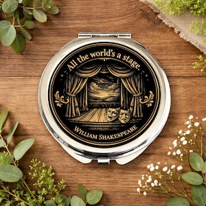 May include: A silver-toned, round compact mirror with a black and gold design. The design features a theater stage with curtains, masks, and the quote "All the world's a stage" by William Shakespeare.