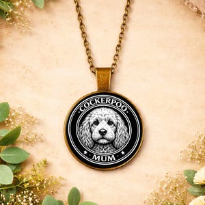 Pode incluir: Colar com pingente redondo com ilustração de um cão Cockerpoo a preto e branco. O pingente tem as palavras "COCKERPOO" e "MUM". A corrente e o pingente são de cor bronze.