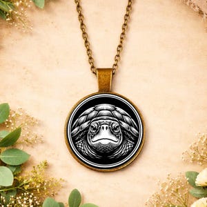 Puede incluir: Un collar con colgante de tono bronce con una ilustración en blanco y negro de una tortuga marina. La cara detallada de la tortuga está dentro de un marco circular, suspendido de una cadena a juego. El colgante tiene una estética vintage.