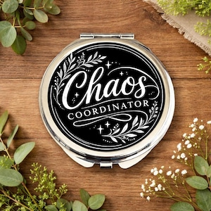 May include: A silver compact mirror with the words "Chaos COORDINATOR" in white script on a black background. The design includes floral and star accents. The mirror is round and sits on a wooden surface.