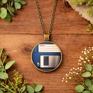 May include: A round pendant necklace featuring a blue and silver floppy disk design. The pendant has a gold-colored frame and chain. The word "INDEX" is printed on the pendant. The necklace is displayed on a wooden surface with greenery.