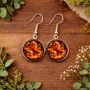 May include: A pair of silver-toned earrings with a circular pendant featuring a fiery horse design. The horse is depicted in shades of red, orange, and yellow, set against a dark background. The earrings have a hook closure.