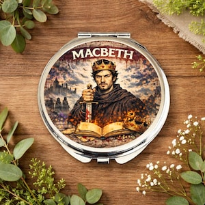 Peut inclure: Un miroir de poche rond et argenté avec une illustration détaillée d'un homme couronné tenant une épée. Le texte "MACBETH" est affiché au-dessus de l'image. L'œuvre comprend un château, un livre ouvert et une couronne.