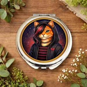 May include: A round, silver compact mirror featuring an orange tabby cat with green eyes, styled in an emo aesthetic. The cat wears a black hoodie, a red and black striped shirt, and a necklace. A broken heart is on the jacket.