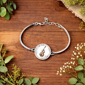 Può includere: Bracciale argentato con un ciondolo circolare con un'illustrazione di violino e note musicali. Il ciondolo è centrato su un bracciale argentato con chiusura a fermaglio. Il bracciale è esposto su una superficie di legno, con vegetazione.