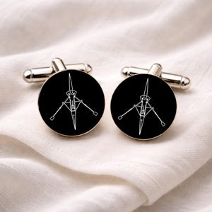 May include: A pair of silver cufflinks with a black circular face. Each cufflink features a white line drawing of a rowing shell. The cufflinks are displayed on a white, textured fabric.