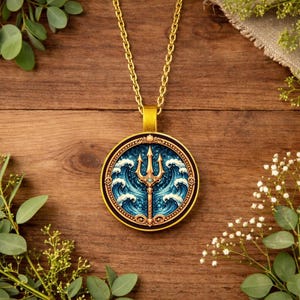 May include: A gold-coloured pendant necklace. The round pendant features a trident design set against a blue background with white waves. The central trident is gold-coloured. The necklace rests on a wooden surface.