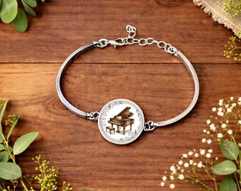 Puede incluir: Pulsera de brazalete plateada con un dije redondo que presenta una ilustración detallada de un piano de cola y un taburete, rodeado de notas musicales. La pulsera se muestra sobre una superficie de madera. El dije mide unos 2,5 cm de diámetro.