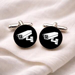 May include: Silver cufflinks with a black circular face, each bearing a white security camera graphic. The cufflinks are presented on a white, textured fabric.
