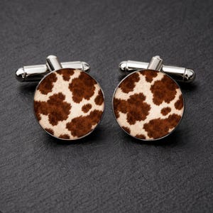 May include: A pair of silver cufflinks with a round face featuring a brown and white cowhide pattern. The cufflinks have a classic design with a t-bar closure.
