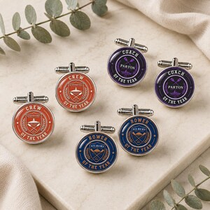 May include: Six round silver-toned cufflinks featuring circular emblems. Two are orange with text reading CREW OF THE YEAR, two are purple with COACH OF THE YEAR, and two are blue with ROWER OF THE YEAR. Each emblem displays crossed oars and laurel leaves.