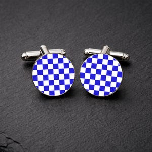 May include: A pair of silver cufflinks with a circular design. Each cufflink has a blue and white chequered pattern, creating a bold and geometric aesthetic. The cufflinks are displayed on a dark, textured surface.