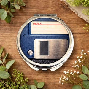 Può includere: Un specchietto compatto rotondo e argentato con un design a floppy disk retrò blu. Lo specchietto presenta la parola "INDEX" su un'etichetta. Il compatto è su una superficie di legno, circondato da verde.