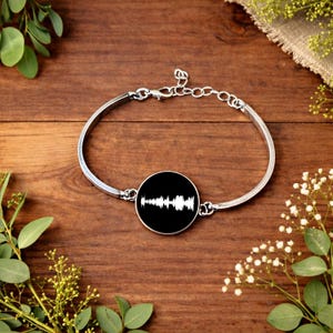 Puede incluir: Pulsera de brazalete plateada con un colgante negro circular. El colgante presenta un diseño de onda de sonido blanco. La pulsera se muestra sobre una superficie de madera con vegetación.