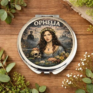 Op de afbeelding: Een ronde, zilveren compacte spiegel met een illustratie van Ophelia uit Hamlet. De afbeelding toont een vrouw in een blauwe jurk die een schedel in het water vasthoudt, met de tekst "Ophelia Hamlet" bovenaan.