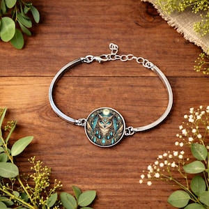 May include: Silver-toned bracelet featuring a circular pendant with an owl design. The owl has blue eyes and teal accents, set against a dark background with a moon. The bracelet is on a wooden surface.