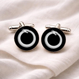 Può includere: Un paio di gemelli argentati con una faccia circolare nera. Ogni faccia presenta un design di ouroboros bianco, un serpente che si morde la coda. I gemelli sono esposti su una superficie bianca strutturata.