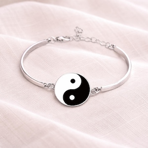 May include: Silver bangle bracelet with a central yin yang symbol. The yin yang design is black and white. The bracelet has an adjustable chain clasp. The bracelet is shown on a white surface.