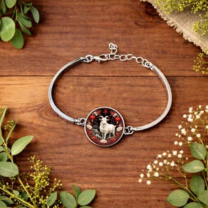 Könnte beinhalten: Silberfarbenes Armreif-Armband mit einem runden Anhänger mit einer weißen Ziegenillustration, roten Laternen und chinesischen Schriftzeichen. Das Armband liegt auf einem Holzgrund mit Grün.