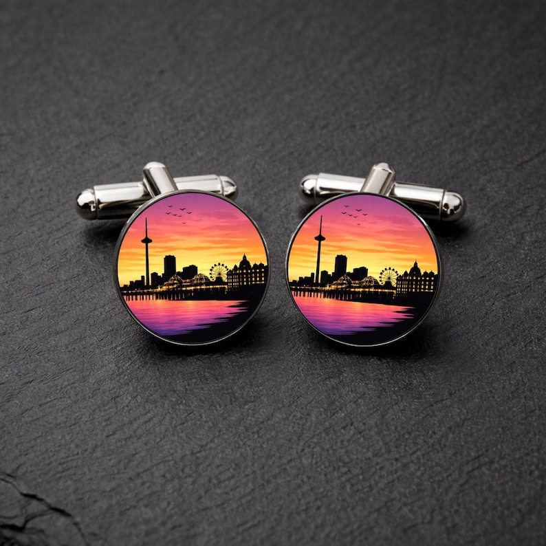 K&ouml;nnte beinhalten: Ein Paar silberne Manschettenkn&ouml;pfe mit kreisf&ouml;rmigem Design. Jeder Manschettenknopf zeigt eine Silhouette einer Stadtlandschaft vor einem Sonnenuntergangshimmel in Orange, Lila und Gelb. Die Manschettenkn&ouml;pfe liegen auf einer dunklen Oberfl&auml;che.
