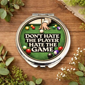 Könnte beinhalten: Ein runder, silberner Kompaktspiegel mit einem grünen Billardtisch-Design. Der Text "DON'T HATE THE PLAYER HATE THE GAME" ist abgebildet. Bunte Billardkugeln und eine Hand, die einen Queue hält, sind ebenfalls zu sehen.