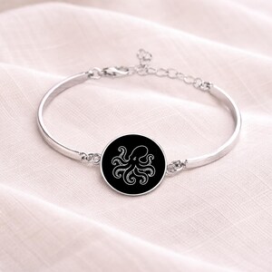 May include: Silver-toned bangle bracelet featuring a black circular charm with a white octopus design. The bracelet has a simple, elegant design and is likely made of metal. The octopus design is a detailed, stylized illustration.