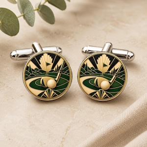 May include: A pair of round silver-tone cufflinks featuring a golf-themed design. The circular face displays a green golf course, a golf club, and a textured golf ball in cream and gold tones, set against a dark background with sunburst rays.