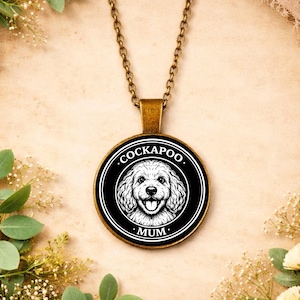 May include: A round pendant necklace with a bronze chain. The pendant features a black and white illustration of a Cockapoo dog with the word "COCKAPOO" at the top and "MUM" at the bottom. The background is a light beige.