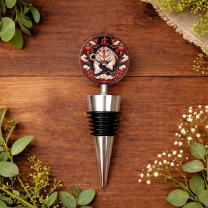 Op de afbeelding: Een decoratieve flesstopper met een zilveren basis en een zwarte rubberen afdichting. De bovenkant heeft een rood cirkelvormig ontwerp met een witte tijgerillustratie en Chinese karakters. Houten achtergrond met groen.