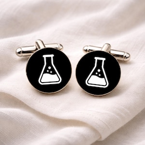 May include: A pair of silver cufflinks with a black circular face. Each cufflink features a white, stylized laboratory flask design. The flask is filled with a white liquid and has three small white dots. The cufflinks are displayed on a white, textured surface.
