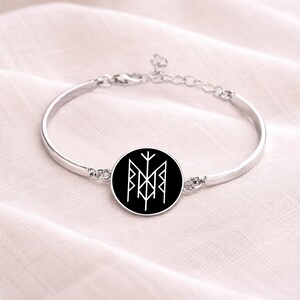 May include: Silver-toned bangle bracelet featuring a black circular charm with a white geometric design. The bracelet has a simple, elegant design and is likely made of metal. The charm's design is a stylized symbol.