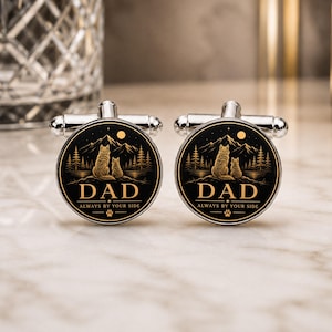 Könnte beinhalten: Ein Paar runde, silberfarbene Manschettenknöpfe mit schwarzer, kreisförmiger Einlage und einer goldfarbenen Grafik von zwei Wölfen vor Bergen und Bäumen. Der Text DAD und ALWAYS BY YOUR SIDE ist in Gold mit einer kleinen Pfote gedruckt.