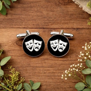 May include: A pair of silver cufflinks with black faces featuring the comedy and tragedy theater masks. The masks are white and set against a black background. The cufflinks are displayed on a wooden surface.