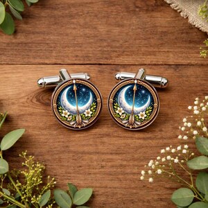May include: Two silver cufflinks with a celestial design. Each cufflink has a crescent moon, arrow, and floral details against a starry backdrop. The cufflinks are round with a gold-coloured border.