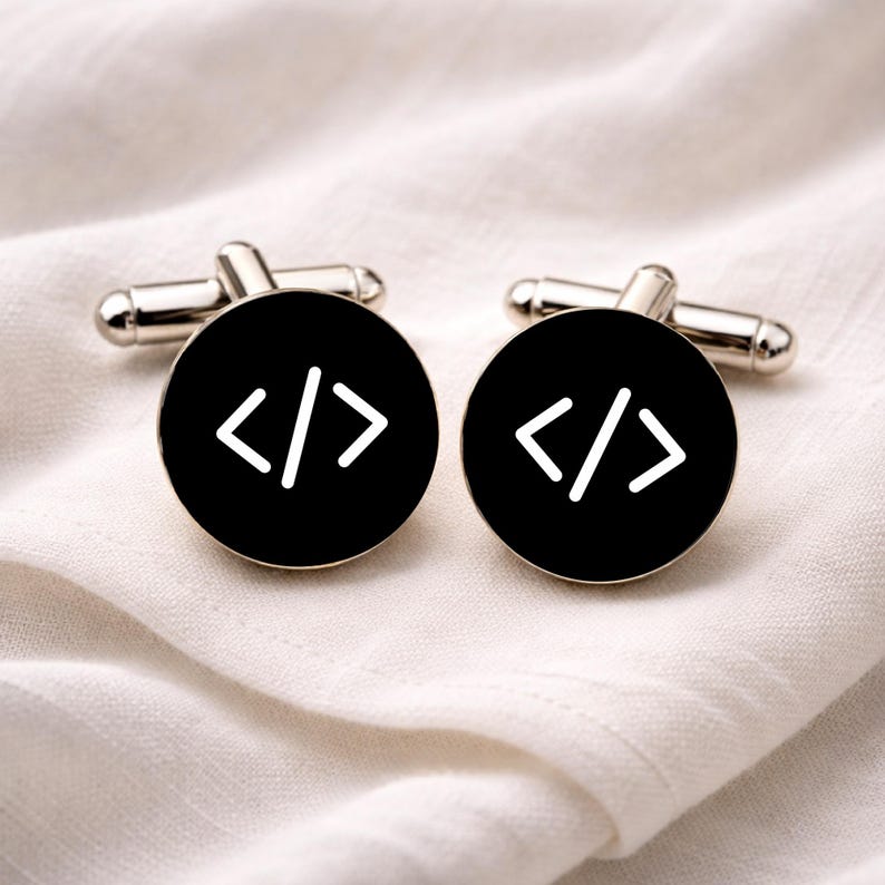 K&ouml;nnte beinhalten: Silberne Manschettenkn&ouml;pfe mit schwarzem, kreisf&ouml;rmigem Gesicht. Jedes Gesicht zeigt ein wei&szlig;es Codesymbol. Die Manschettenkn&ouml;pfe werden auf einem wei&szlig;en, strukturierten Stoff pr&auml;sentiert.