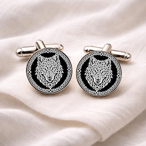 Könnte beinhalten: Zwei silberne Manschettenknöpfe mit einem schwarzen, kreisförmigen Gesicht, das ein weißes Wolfsdesign zeigt. Der Wolf ist in einem komplizierten, keltisch inspirierten Stil dargestellt. Die Manschettenknöpfe liegen auf weißem, strukturiertem Stoff.