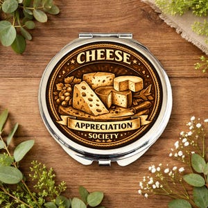 Könnte beinhalten: Ein runder, silberner Kompaktspiegel mit einem braunen und goldenen Design, das verschiedene Käsesorten und den Text "CHEESE APPRECIATION SOCIETY" zeigt. Der Spiegel ist geöffnet und zeigt die reflektierende Oberfläche. Auf einem Holzgrund mit Grünzeug.