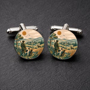 May include: A pair of round silver-tone cufflinks featuring a printed illustration of a person in a green jacket standing in a hilly landscape with two dogs and a deer. The scene includes a sun, trees, and a small village in the distance.