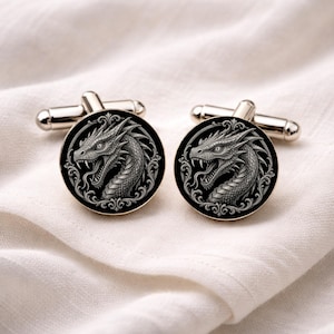 Könnte beinhalten: Ein Paar silberne Manschettenknöpfe mit schwarzem, kreisförmigem Gesicht, das ein detailliertes Drachendesign zeigt. Der Drache ist in Graustufen dargestellt, mit komplizierten Schuppen und einem bedrohlichen Ausdruck. Klassisches, elegantes Design.