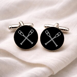 May include: A pair of silver cufflinks with a black circular face. Each face features a white outline of two crossed oars. The cufflinks are displayed on a white, textured surface.