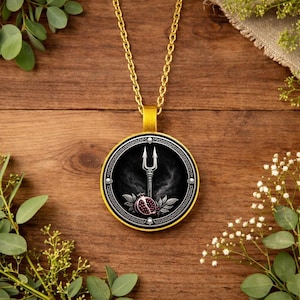 Può includere: Collana con pendente dorato con un pendente rotondo. Il pendente ha uno sfondo nero con un tridente argentato, un melograno ed elementi decorativi. La collana è esposta su una superficie di legno.