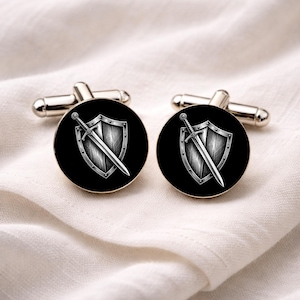 May include: A pair of silver cufflinks with a black circular face. Each cufflink features a detailed illustration of a sword and shield in a silver color. The cufflinks are displayed on a white fabric background.