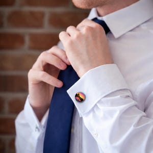 K&ouml;nnte beinhalten: Runde Manschettenkn&ouml;pfe mit einem Sonnenuntergangsdesign in Orange, Rosa und Schwarz. Die Manschettenkn&ouml;pfe werden auf einem wei&szlig;en Hemd mit einer blauen Krawatte getragen. Der Hintergrund ist eine Backsteinmauer.