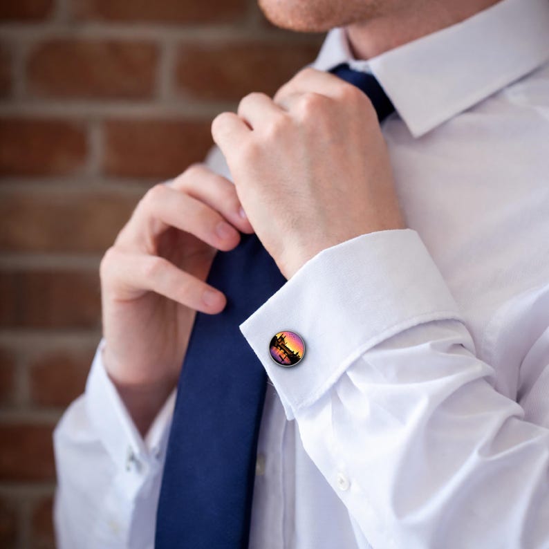 K&ouml;nnte beinhalten: Runde Manschettenkn&ouml;pfe mit einem Sonnenuntergangsdesign in Orange, Rosa und Schwarz. Die Manschettenkn&ouml;pfe werden auf einem wei&szlig;en Hemd mit einer blauen Krawatte getragen. Der Hintergrund ist eine Backsteinmauer.