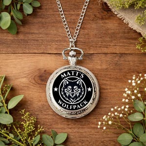 May include: A silver pocket watch on a chain, with a black face displaying a white wolf design and the words "MATT'S WOLFPACK". The watch is set against a wooden background with foliage.